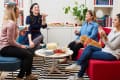 Four women enjoying snacks and drinks in a cozy living room with a small dog on a red chair.