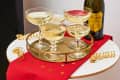 Champagne glasses on a gold tray with a bottle of prosecco on a red cloth with gold stars.
