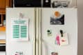 Refrigerator with $100 savings challenge chart, postcards, and magnets, including a pizza slice magnet and a Boston postcard.