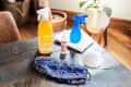 Cleaning supplies on a wooden table, including disinfectant spray, hand sanitizer, blue spray bottle, mask, and cloth.