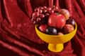 Yellow bowl with pomegranates, plums, and grapes on a red velvet fabric.