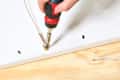 A person using a power drill to secure a brass hook into a white wooden frame, with a wooden surface below.