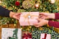 Two hands exchanging a pink gift box with gold ribbon in front of a decorated Christmas tree.