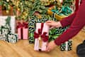 A person placing a pink gift box with a red bow among various decorated presents under a Christmas tree.