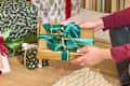 A person holding a gold-wrapped gift with a teal ribbon, surrounded by various decorated presents on a wooden floor.
