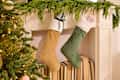 Stockings hanging from a mantel decorated with greenery, alongside a Christmas tree and stacked books.