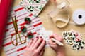 A hand writing a gift tag on a wooden table, surrounded by wrapping paper, scissors, mini wreaths, and decorative ribbon.