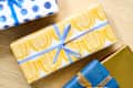 Three beautifully wrapped gifts on a wooden surface: one with blue polka dots, one with yellow patterned paper, and one in blue with an orange ribbon.