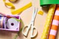Crafting supplies on a table, including scissors, colorful ribbons, and patterned wrapping paper rolls.