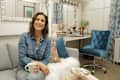 Woman in denim shirt holding a mug, sitting on a gray sofa in a cozy room with blue velvet chair and decorative items.