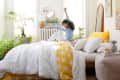 Woman stretching on a bed with white and yellow bedding, surrounded by plants and wall art in a bright bedroom.