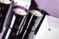 Three lit candles in dark glass holders with white boxes labeled "Beauty Pie" on a purple surface.