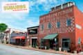 Historic brick building with "Hay Grain Flour & Feed Mills" sign, shops below, and colorful storefronts on a small-town street.