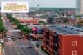 Aerial view of a busy suburban street with red brick buildings, trees, and cars, featuring a "Coolest Suburbs" sign.