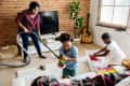 Woman vacuuming living room with two children playing, one with a toy stacker and the other with a box of toys.