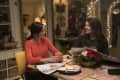 Two women sitting at a kitchen table with newspapers, a mug, and a floral centerpiece, smiling and talking.