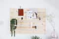 Wooden wall organizer with hanging plants, notebooks, and art prints, next to a pink vase with white flowers.