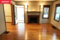 Empty living room with hardwood floors, a brick fireplace, and two windows. Open door leads to a sunlit room.