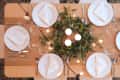 Table set for six with white plates, folded napkins, cutlery, and a centerpiece of greenery and candles.