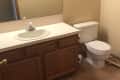 Small bathroom with beige walls, wooden vanity, white sink, and toilet. Brown carpet flooring and large mirror above sink.