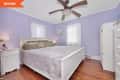 Lavender bedroom with white bed, green pillows, ceiling fan, and wooden floor. White nightstand and wardrobe.