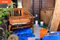 Cluttered patio with wooden bench, orange bucket, blue tarp, and garden tools against a brick wall and wooden fence.