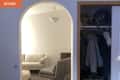 Arched entryway to living room with beige sofa, gray chair, and pouf; adjacent open closet with hanging clothes.