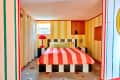 Striped bedroom with a pink and orange bedspread, checkered bed frame, red lamp, and yellow striped walls.