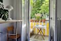 Bright balcony view with potted plants, a small table set for two, and a wooden chair beside a white lamp.