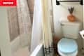 Small bathroom with a shower, white curtains, a blue toilet seat, and an orange vase with dried flowers.