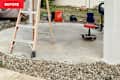 A wooden ladder stands on a concrete surface surrounded by gravel, with a red rolling stool and various buckets nearby.