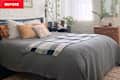 Cozy bedroom with a gray bedspread, colorful pillows, a plaid throw, and a stack of books beside a wooden nightstand.