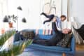 Man lying on blue sofa lifting a child in striped shirt, surrounded by modern decor and plants.