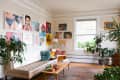 Living room with a beige tufted bench, colorful art on walls, wooden coffee table, and potted plants by a large window.