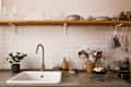 Kitchen sink with potted plant, utensils in a jug, and a wooden shelf with dishes against a white tiled backsplash.