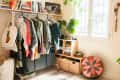 Clothing rack with colorful clothes, books on a shelf, potted plant, and woven baskets in a bright room.