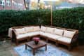 Outdoor patio with wooden sectional sofa, cream cushions, blue patterned rug, and potted flowers under string lights.