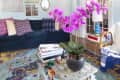 Living room with blue sofa, colorful pillows, glass coffee table with books, and pink orchids, against a gallery wall.