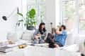 Couple sitting on a white sofa with a cat and dog, surrounded by plants and large windows.