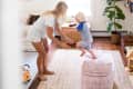 Woman playing with child in a bright living room with a large plant, bookshelf, and patterned ottoman.
