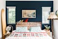 Bedroom with rustic wooden headboard, surfboard, vintage lamp, and patterned quilt against a dark blue wall.