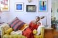 Woman in red dress on yellow sofa with colorful pillows, surrounded by eclectic wall art and a gold lamp.
