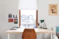 Home office with wooden desk, green lamp, chair, wall art, and window view of houses.