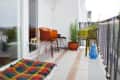 Small balcony with colorful chairs, striped rug, potted plants, and a blue vase, overlooking a cityscape.