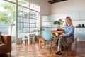 Woman sitting at a wooden dining table in a bright kitchen with large windows, plants, and light blue cabinets.