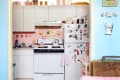 Small kitchen with pink accents, white cabinets, fridge covered in magnets, and a stove with pink pots.