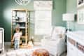Child reaching for toys on a wooden shelf in a green nursery with a white armchair, lamp, and changing table.