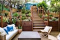 Outdoor patio with wooden steps, string lights, white sofa, blue pillows, and lush greenery.