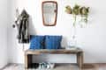 Entryway with wooden bench, blue patterned cushions, hanging plants, mirror, coat hooks, and shoes on a rug.