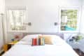 Bright bedroom with a white bed, colorful pillows, wall lamps, and two windows overlooking greenery.